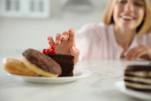 Concept of choice between healthy and junk food. Woman with sweets at white table in kitchen, focus on hand