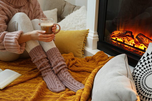 Женщина с чашкой кофе сидит возле горящего камина, крупным планом