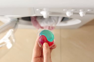 Çamaşır makinesinin yanında çamaşır deterjanı tutan bir kadın var.