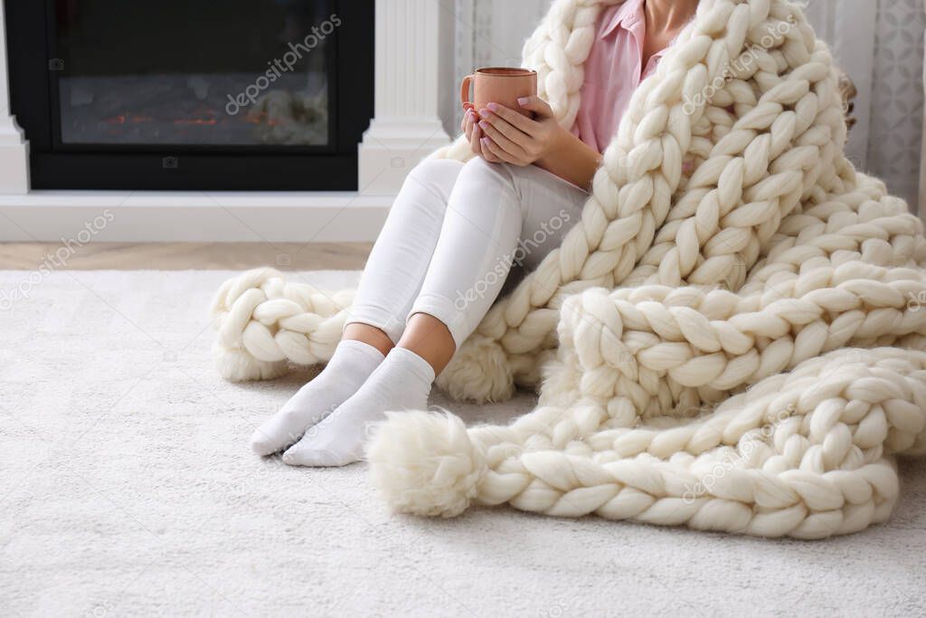 Woman covered with knitted plaid holding cup of hot drink on floor at home, closeup