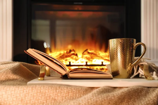 Cup of hot drink and book on blanket near fireplace indoors. Magic atmosphere