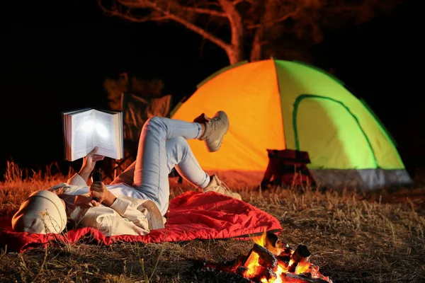 Young woman with flashlight reading book near bonfire at night. Camping season