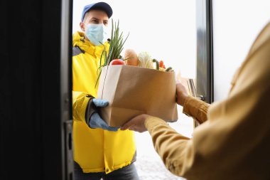 Tıbbi maskeli kurye, kapıdaki kadına poşet dolusu erzak veriyor. Covid-19 karantinası sırasında teslimat servisi