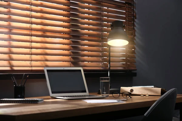 Modern laptop and lamp on table near window in office