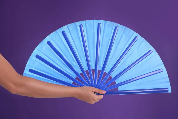 Woman holding blue hand fan on purple background, closeup