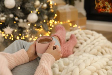 Elinde bir bardak sıcak içecek olan kadın Noel için dekore edilmiş oturma odasında şöminenin yanında dinleniyor.