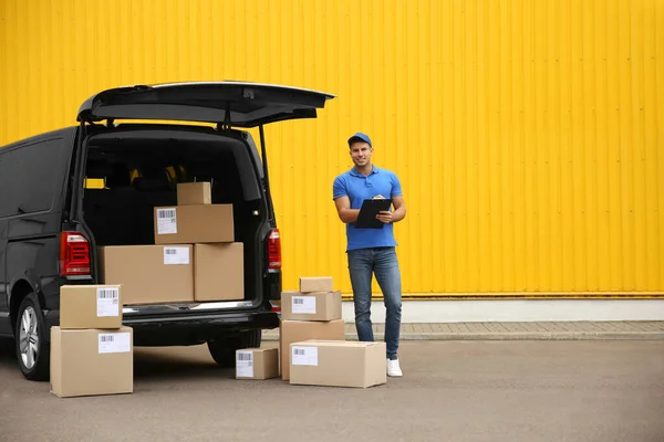 Courier with clipboard and parcels near delivery van outdoors