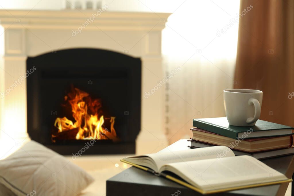 Cup of hot drink and book on table near fireplace at home. Cozy atmosphere