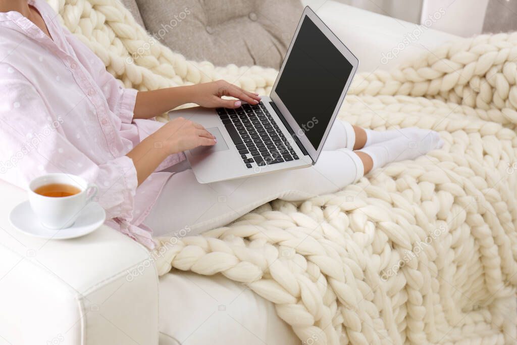 Woman using laptop on couch with soft knitted blanket at home, closeup