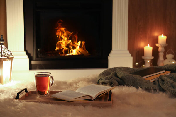 Cup of tea and book on fuzzy rug near fireplace at home. Cozy atmosphere