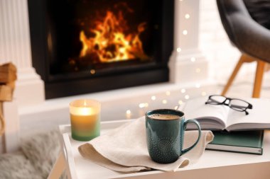Cup of coffee, burning candle and books on tray near fireplace indoors. Cozy atmosphere clipart