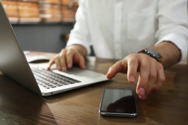 Serbest Çalışan, bilgisayarını kapalı bir masada çalışırken yanına akıllı telefon alıyor.