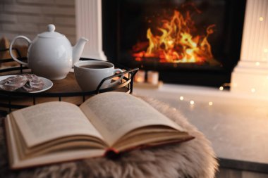 Cup of tea, cookies and book on faux fur near fireplace indoors, space for text. Cozy atmosphere clipart