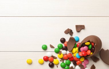 Kırık çikolata yumurtası ve beyaz ahşap masa üzerinde renkli şekerler. Metin için boşluk