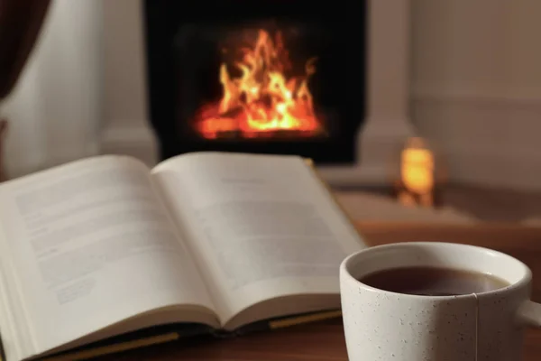 Cup of hot tea and book on wooden table near fireplace at home. Cozy atmosphere