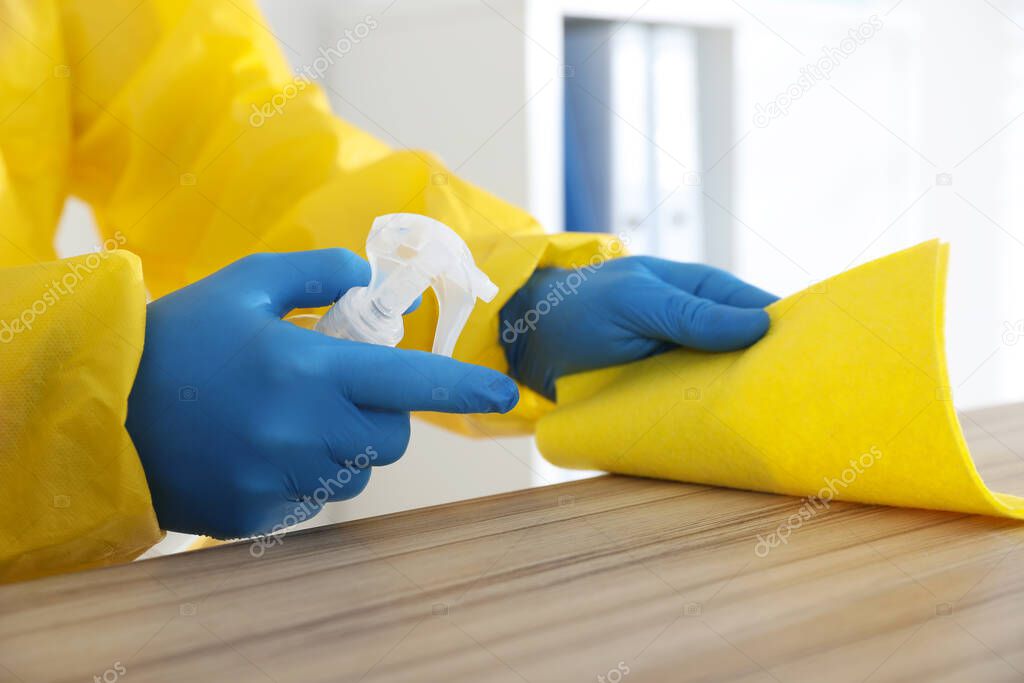 Employee in protective suit and gloves sanitizing wooden countertop indoors, closeup. Medical disinfection