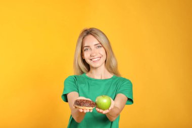 Sarı arka planda donut ve sağlıklı elma arasında seçim yapan bir kadın.