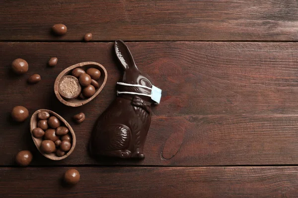 Chocolate bunny with protective mask, eggs and space for text on wooden table, flat lay. Easter holiday during COVID-19 quarantine