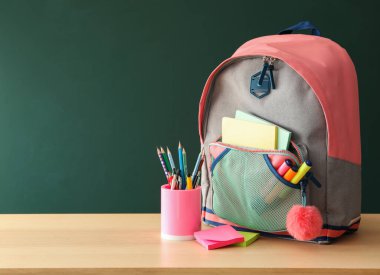 Zarif bir sırt çantası ve yeşil karatahtanın yanındaki ahşap masa üzerinde farklı okul kırtasiyesi. Okula geri dön.