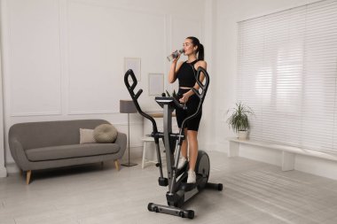 Evdeki eliptik makinede eğitim sırasında su içen mutlu genç kadın.