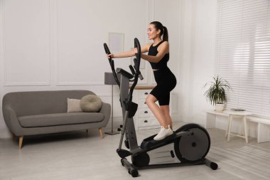 Happy young woman training on elliptical machine at home clipart