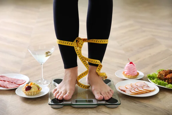 Food, alcohol left after holidays and woman with measuring tape standing on scales indoors, closeup. Overweight problem