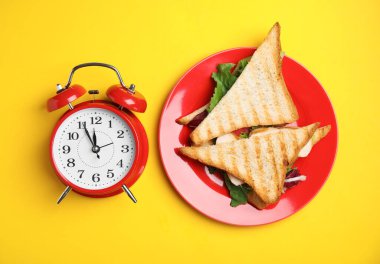 Lezzetli sandviçler ve sarı arka planda çalar saat, düz yatış. Beslenme rejimi