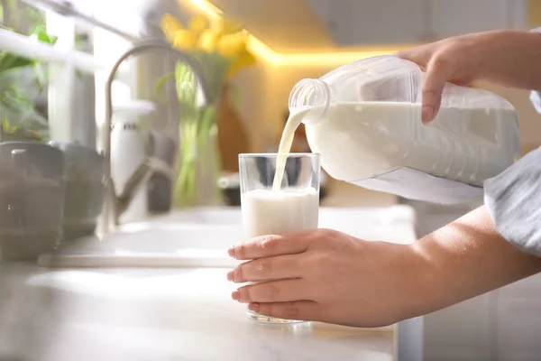 Young woman pouring milk from gallon bottle into glass at white countertop in kitchen, closeup