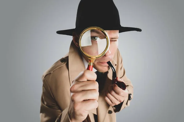 Male detective with smoking pipe looking through magnifying glass on grey background, closeup