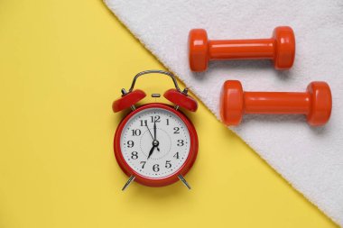 Alarm saati, havlu ve sarı arka plandaki dambıllar düz. Sabah egzersizi