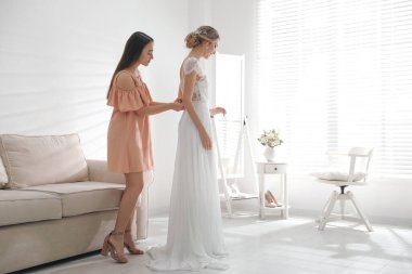 Young woman helping bride to put on wedding dress in room. Space for text