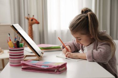 Adorable little girl doing homework with tablet at table indoors