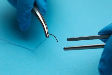 Açık mavi arka planda dikişli forseps tutan profesyonel cerrah, yakın plan. Tıbbi ekipman