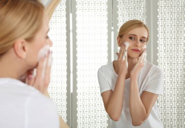 Mutlu genç bir kadın banyoda aynanın yanındaki yüzün üzerine köpük sürüyor.