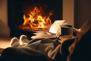 Woman with cup of drink and book near fireplace at home, closeup clipart