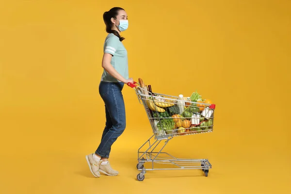 Woman with protective mask and shopping cart full of groceries on yellow background