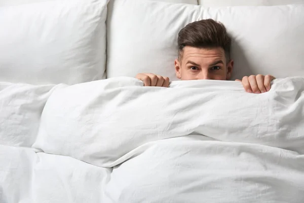 Handsome man covering his face with blanket while lying in bed at home