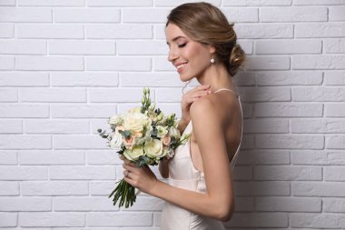 Genç gelin zarif saç stiliyle beyaz tuğla duvarın yanında düğün buketi tutuyor.