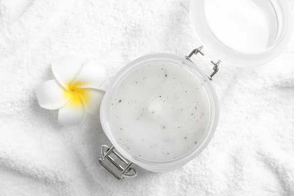 Jar of salt scrub and plumeria on white towel, flat lay