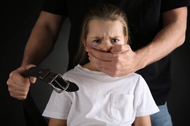 Siyah arka planda küçük kızın ağzını kapatan kemerli bir adam. Aile içi şiddet