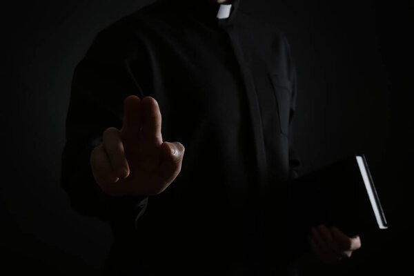 Priest with Bible making blessing gesture on dark background, closeup