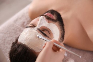 Kozmetik uzmanı spa salonunda adamın yüzüne maske takıyor.
