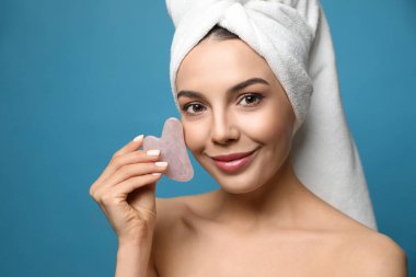 Güzel genç bir kadın mavi arka planda gua sha aracıyla yüz masajı yapıyor.