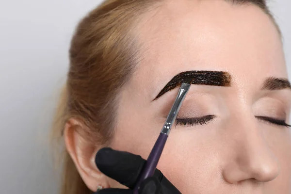 Beautician applying tint during eyebrows correction procedure on light grey background, closeup