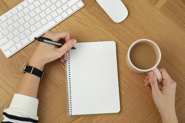 Left-handed woman with cup of coffee writing in notebook at wooden table, top view