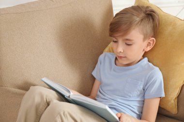 Evdeki koltukta kitap okuyan küçük çocuk.