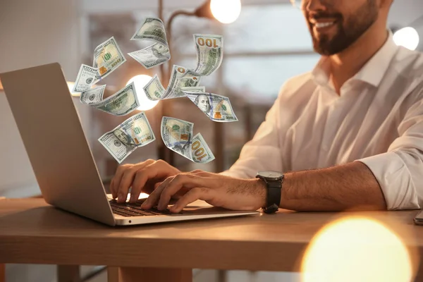 Making money online. Closeup view of man using laptop at table and flying dollars