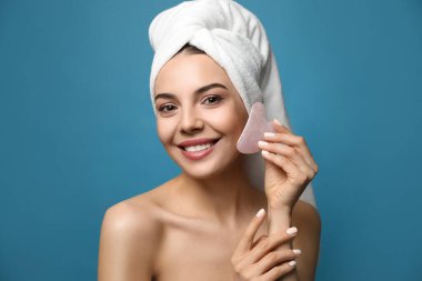 Güzel genç bir kadın mavi arka planda gua sha aracıyla yüz masajı yapıyor.
