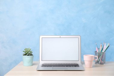 Boş ekranlı modern laptop, açık mavi duvarın yanındaki beyaz masada fincan ve ofis malzemeleri. Metin için boşluk