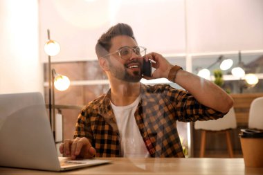 Kafede dizüstü bilgisayarla çalışan bir adam akıllı telefondan konuşuyor.